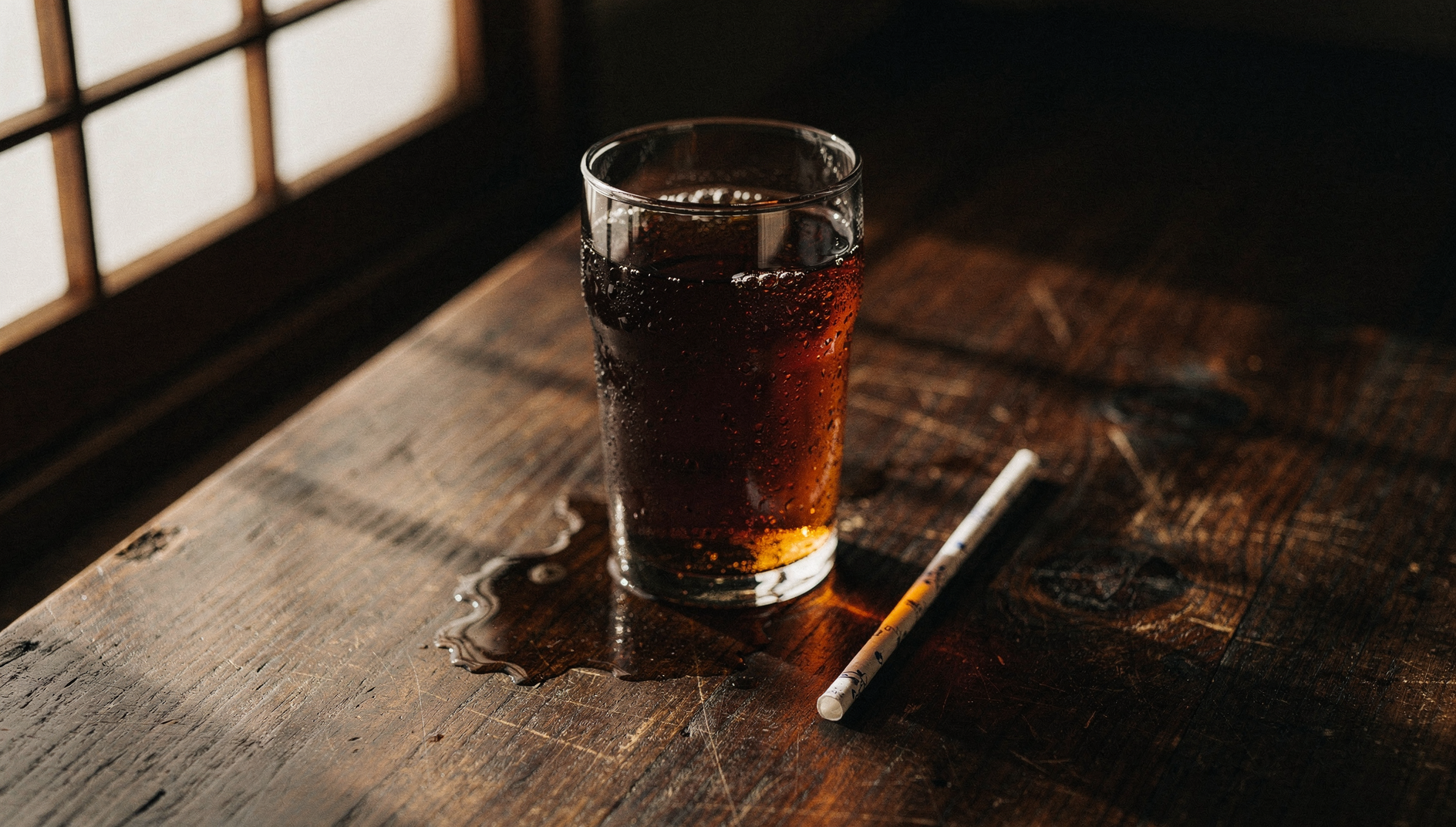 Hohes Glas mit dunkler Cola und Kondenstropfen auf einem alten Eichentisch, weiches Spätnachmittagslicht aus einem Sprossenfenster (KI-generiert)
